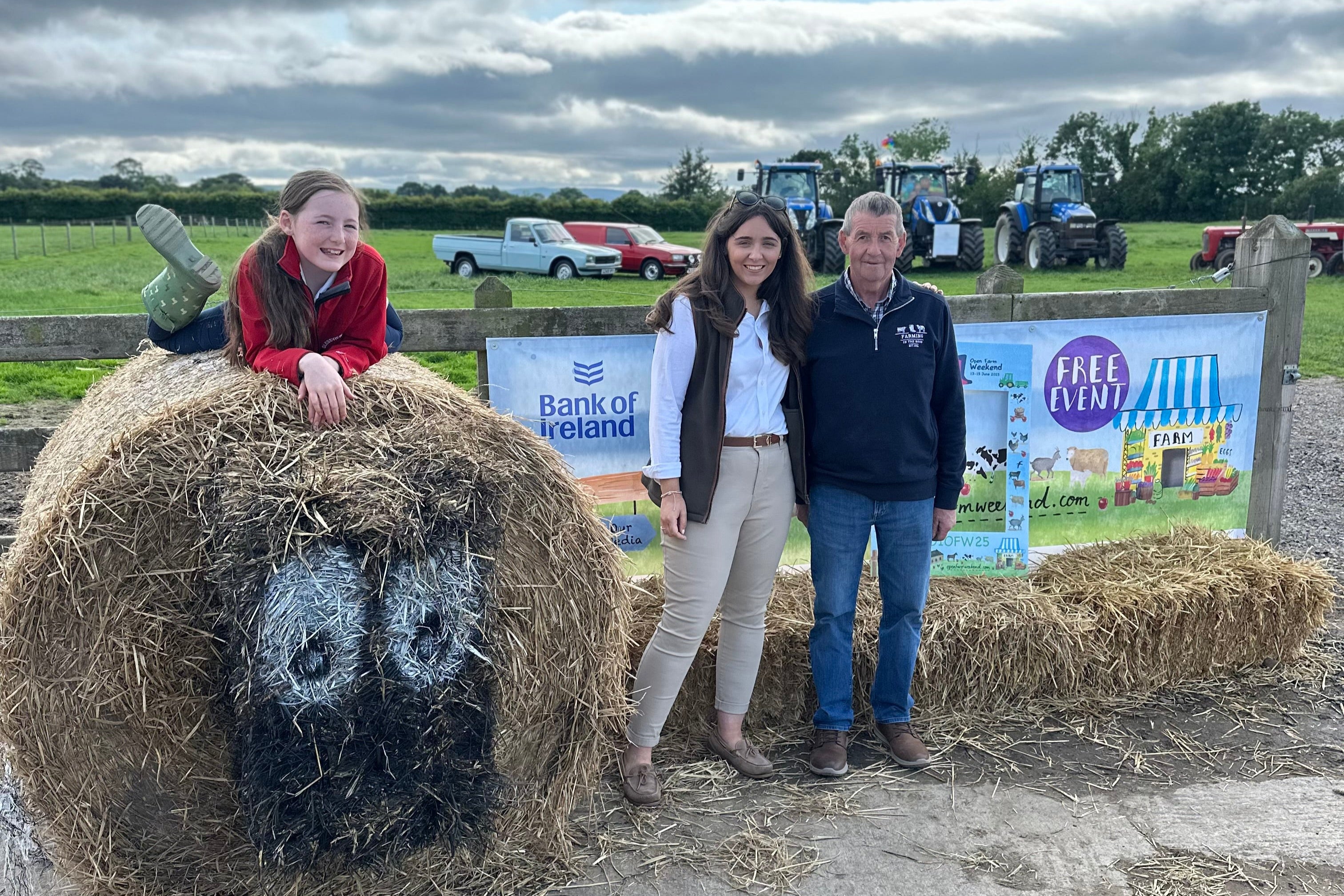 People posing with a hay bale at an agricultural event with tractors and signs in the background.