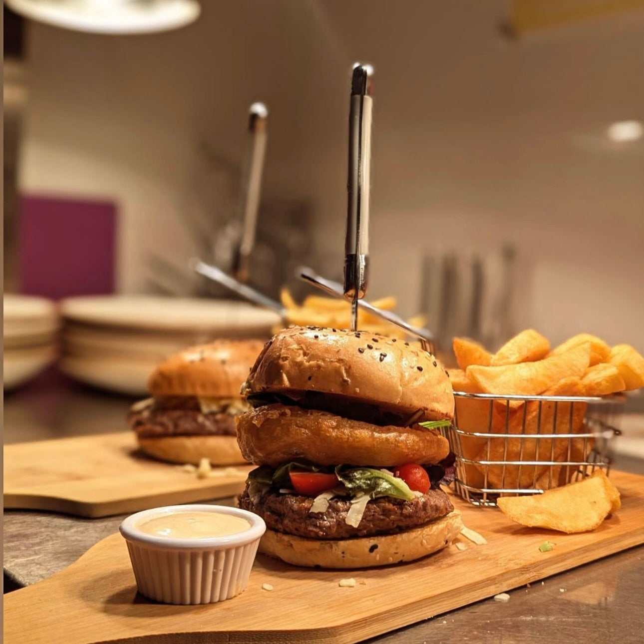 Burger on a wooden board with fries and a side of sauce, set against a blurred kitchen background.