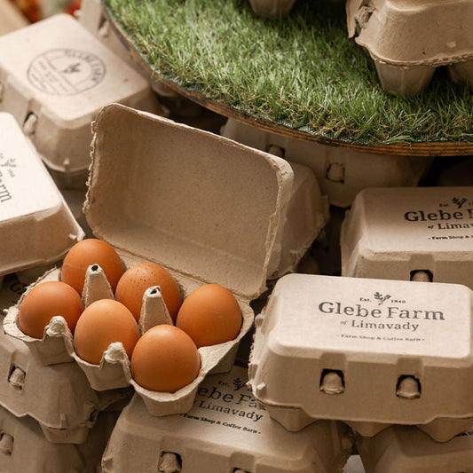 A close-up of stacked paper egg cartons branded 'Glebe Farm + Limavady' with several brown eggs visible in one open carton, set against a rustic background with grass and wood.